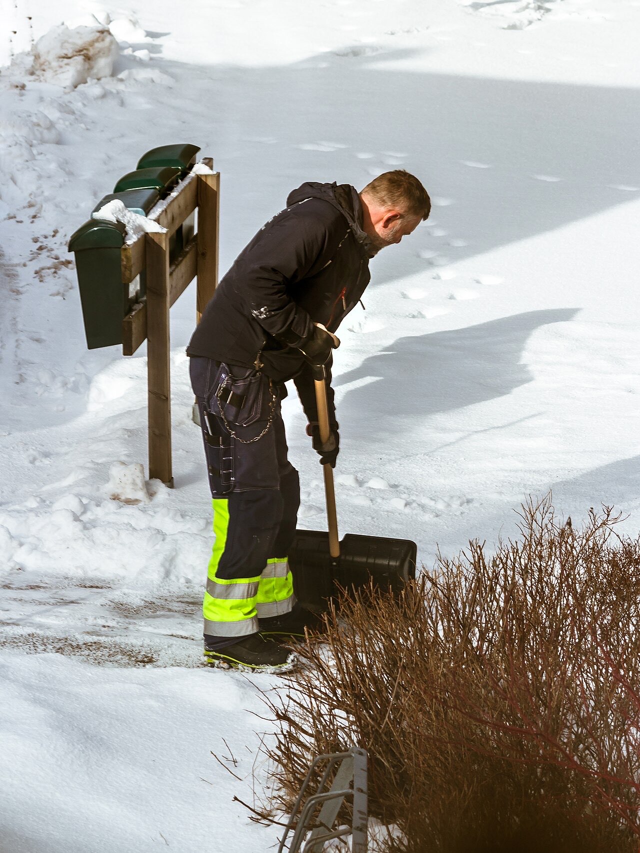 A man in work clothes shoveling snow beside a row of mailboxes